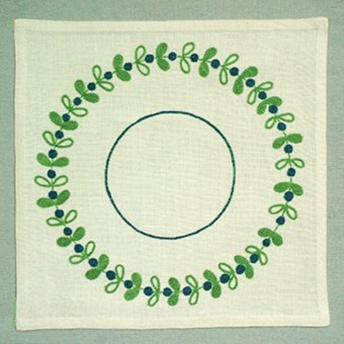 Duk broderad med blå bär och gröna blad i krans. Blå ring i mitten.