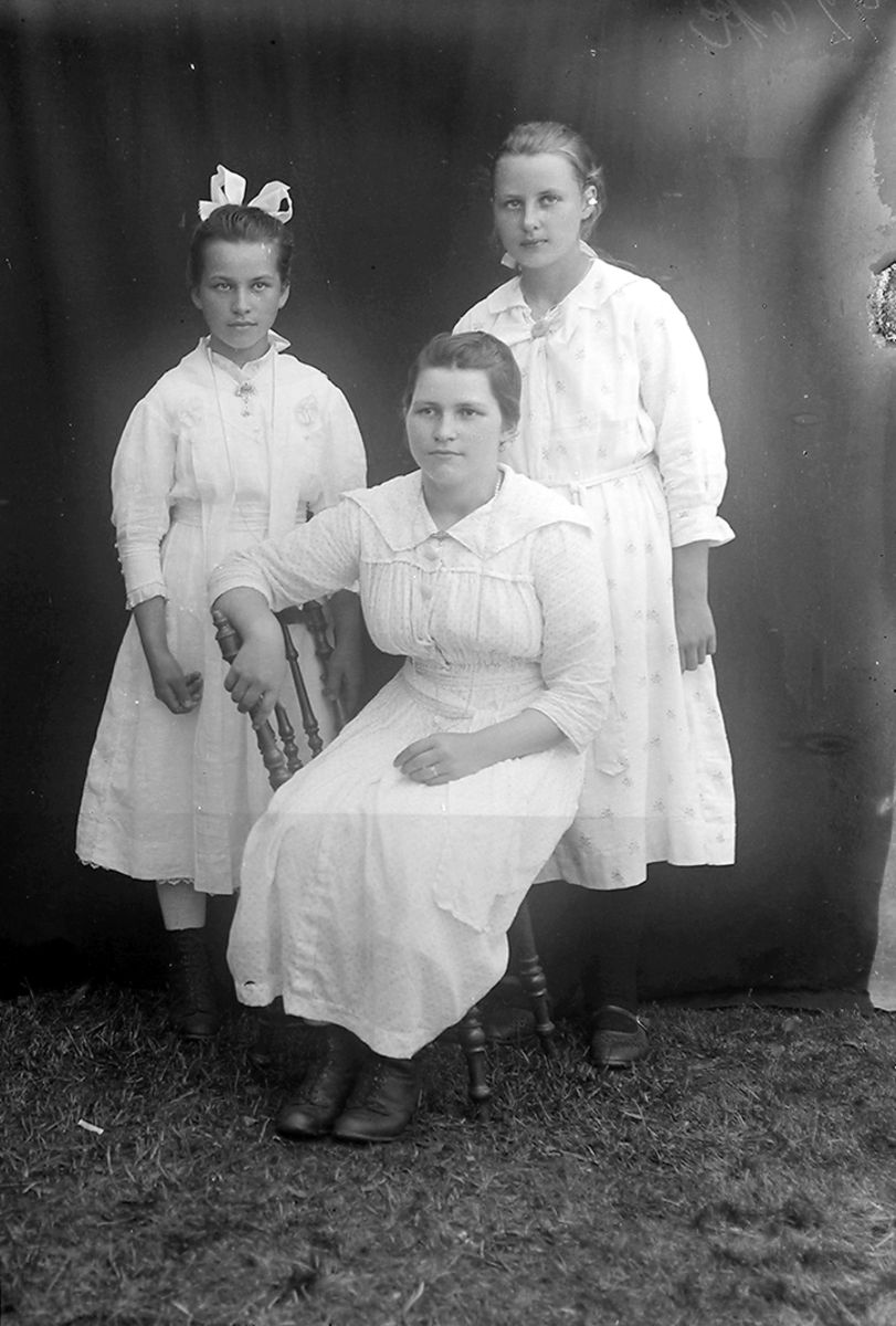Portrett. Tre jenter. En jente sitter på en stol, to står.Bildet er tatt i Sørfold.Muligens konfirmanter i 1919.