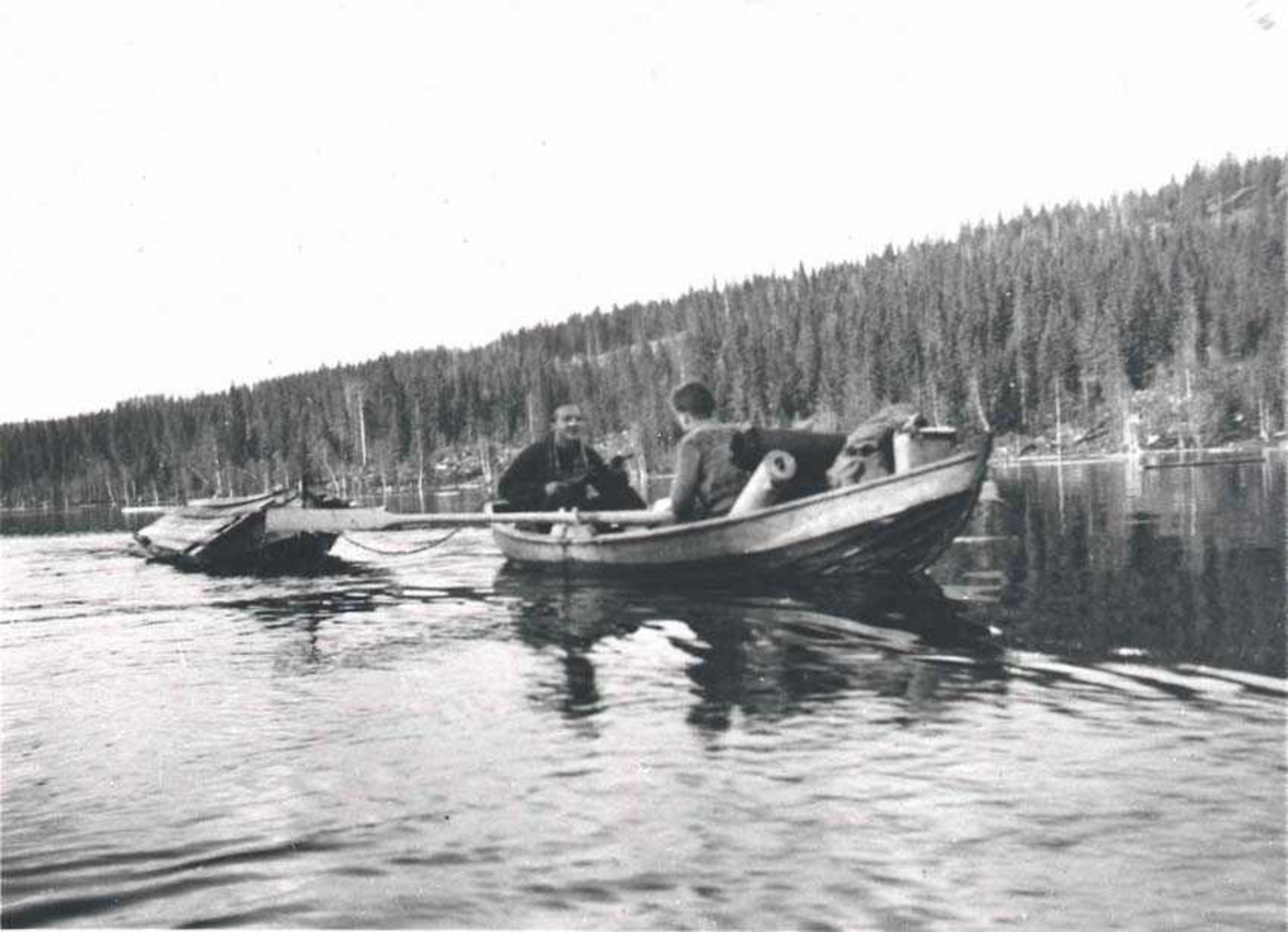To personer i en robåt ute på et vann. En båt på slepp. Skog i bakgrunnen.