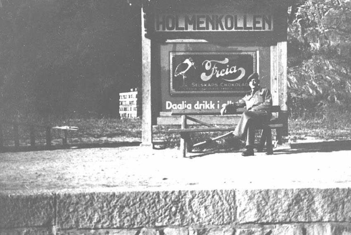 En person som sitter på en benk ved et skilt. Mann i uniform. På skiltet står det Holmenkollen øverst med reklame for Freia under. 