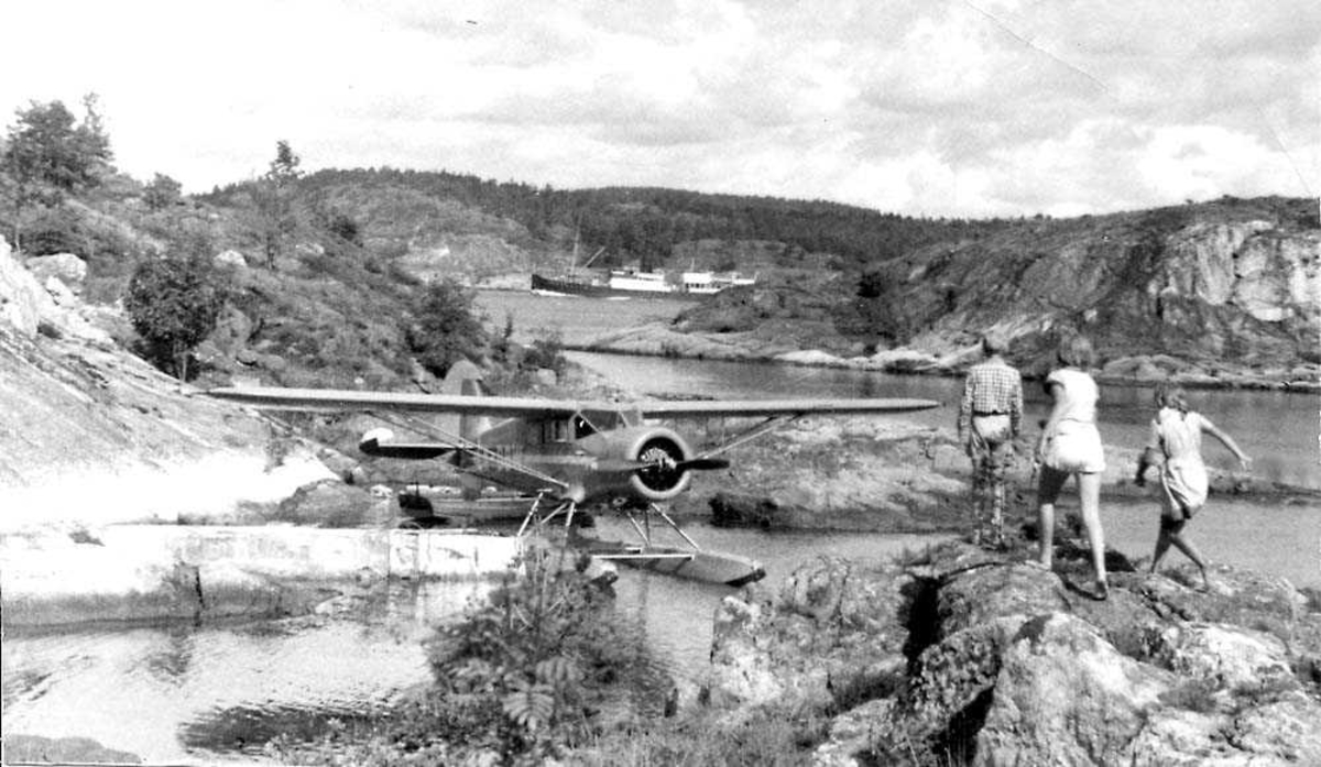 Ett fly som ligger ved en kai, Polar C-5 med flotørrer LN-DBW fra Widerøes Flyveselskap A/S. Tre personer i forgrunnen. En båt og fjell i bakgrunnen.