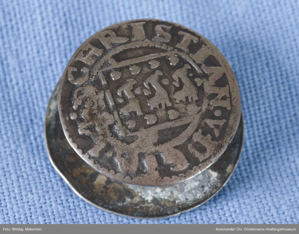 Laget av mynter som er preget m/ monogram, satt sammen til en knapp.