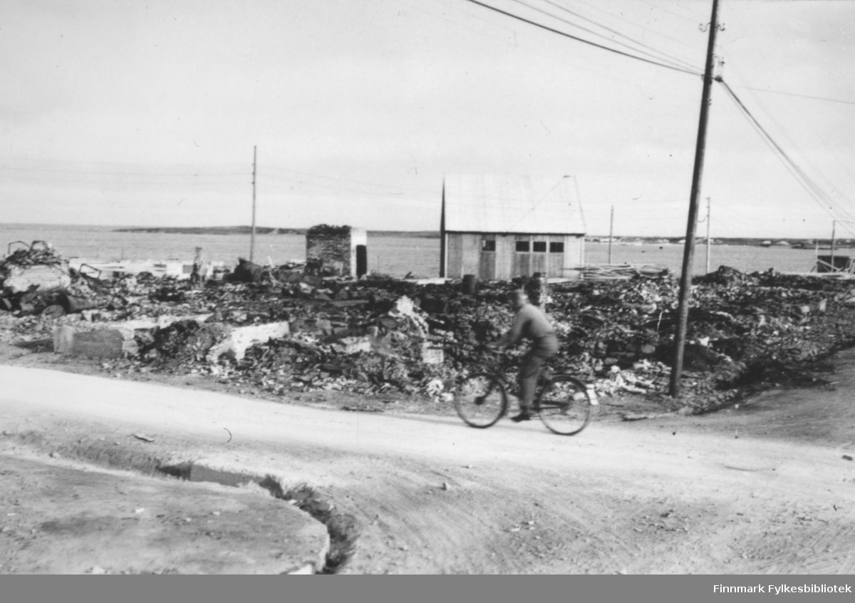 Ruiner i Vadsø sentrum etter andre verdenskrig. I forgrunnen en vei og en mann som sykler der. Til høyre for ham står det en stolpe. I bakgrunnen er det en tomt med ruiner, en liten bygning, og bak der igjen ser man havet