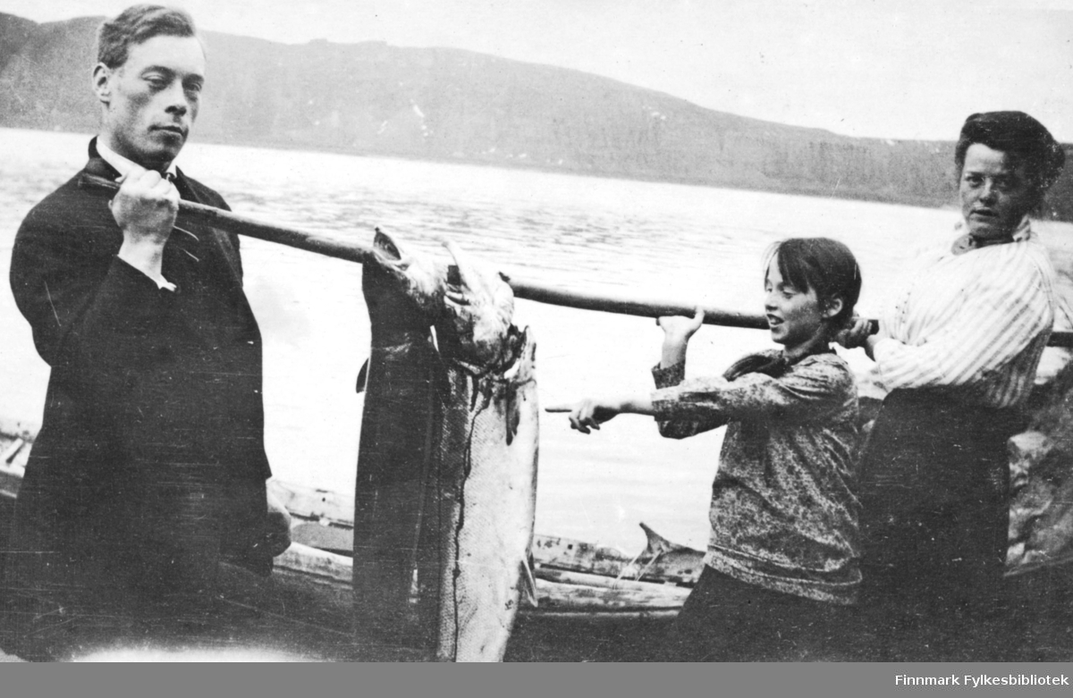 Stolte fiskere viser frem sin fangst. En mann, en dame og en jente viser frem to flotte laks på henholdsvis 11 og 12 kg. Antakelig på Langnes i Tana. Bak dem ligger det en elvebåt. I bakgrunnen ser vi Tanafjorden.