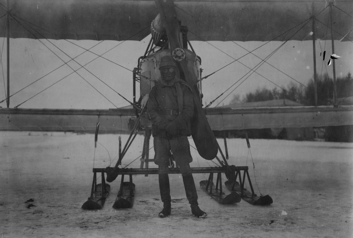 Porträtt. En militär flygare med flygarglasögon ansiktet täckt för kylan står framför flygplan Albatros BIIa med skidor på ett flygfält, vintertid. I bakgrunden syns tälthangarer.