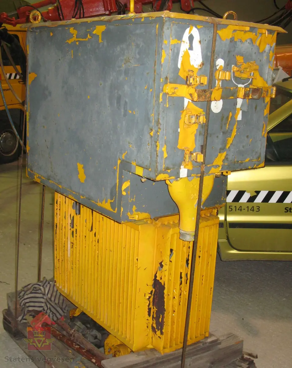 Gul tre-faset transformator av metall. Står på fire hjul. Leverer ca. 4000 volt og 50 KVA. Mye av fargen er borte. Skilt fra produsent med fabrikkspesifikasjoner og advarsler.