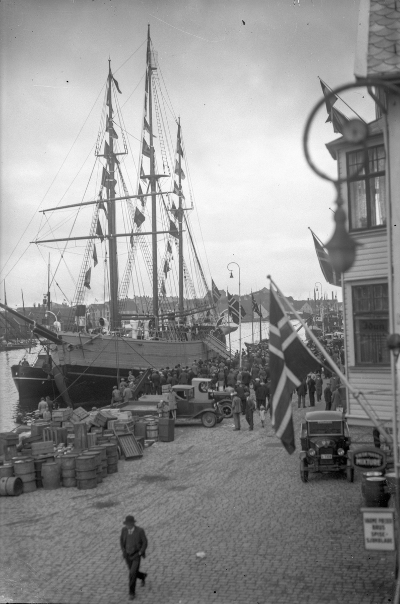 Tremastet skonnert ankret opp ved havn ved Smedasundet i Haugesund. En person i forgrunnen, og mange ved kai ved siden av skipet. Norsk flagg og en bil ved siden av et trehus.