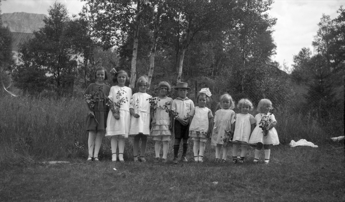 9 småbarn. 8 jenter og en gutt. Jentene står på en rekke, og gutten står i midten med en hatt på hodet.  Trær,et hus og fjell i bakgrunnen.