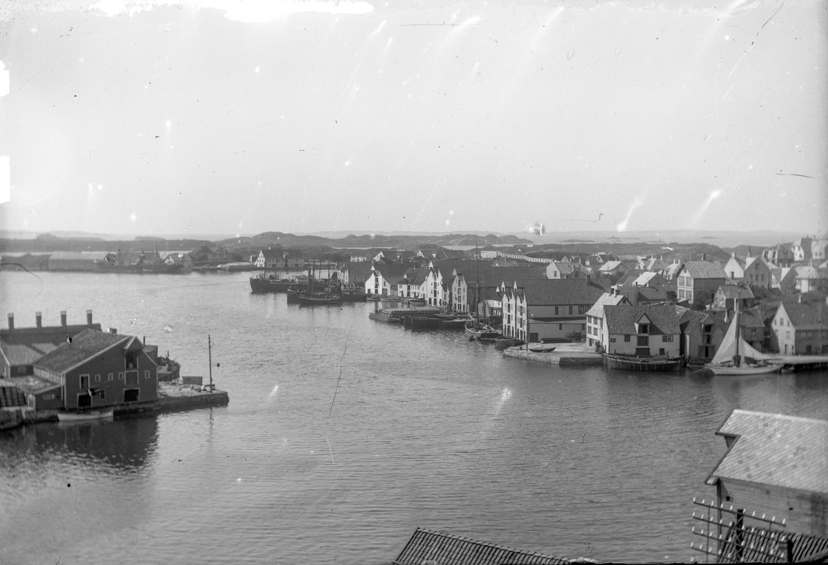 Innseilingen ved Kortenes. Krossen midt på bildet. Risøy til høyre, med flere seilskuter og dampskip ankret opp. Til venstre på bildet en fiskebåt. Sjøhus til venstre og til høyre på bildet. Tønnelaget på en av kaiene på Risøy midt på bildet.