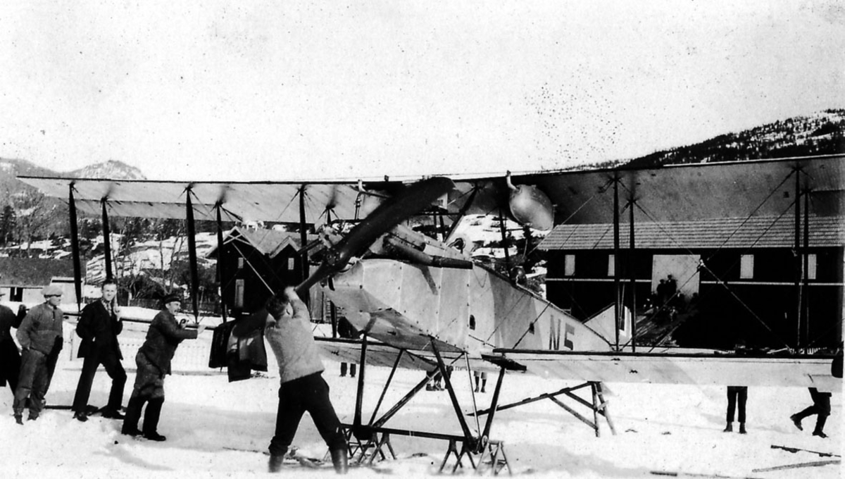 Åpen plass, flere personer ved ett fly på bakken, N5 Avro 501 A (503), med skiunderstell. Bygninger bak, snø på bakken. en person drar i propellen.
