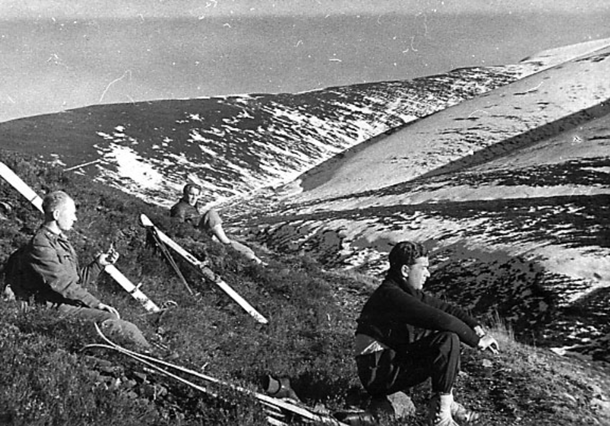 Portrett, tre personer sitter i fjellskråning. Skiutstyr ligger på bakken ved siden av.