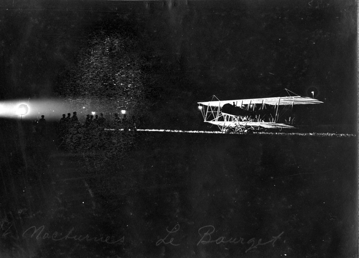 Fly, Maurice Farman. Står på bakken. Skrått forfra. Noen personer foran flyet. Bildet tatt i mørke, hvor flyet er flombelyst.