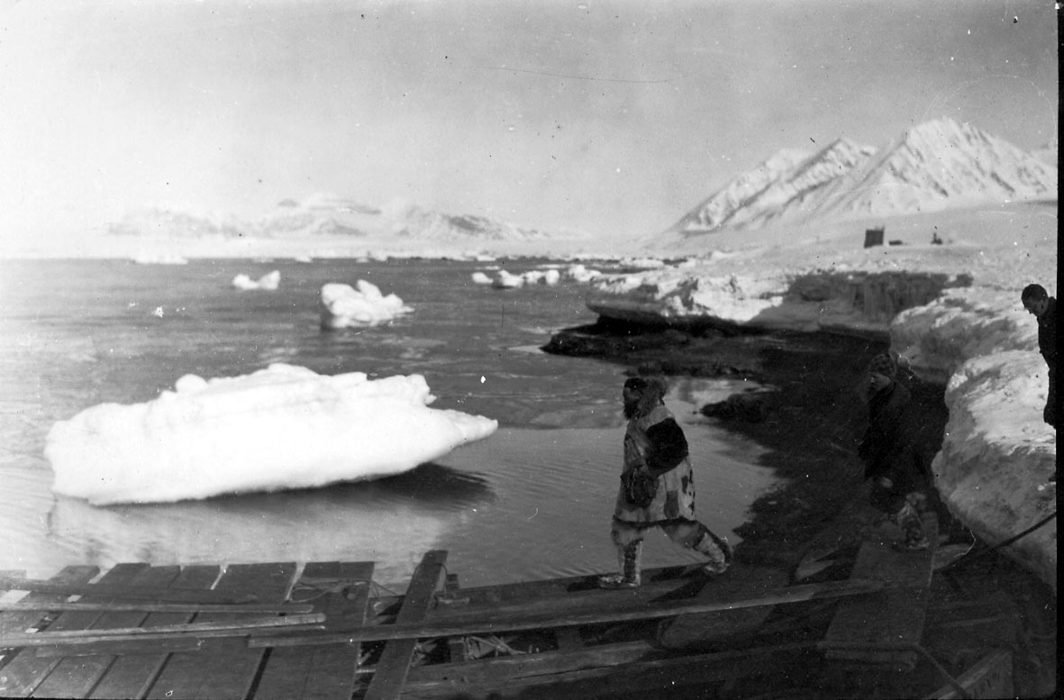 En person på "provisorisk " kai. Mindre isflak i bakgrunnen, hav og fjell. 2 andre personer sees også.