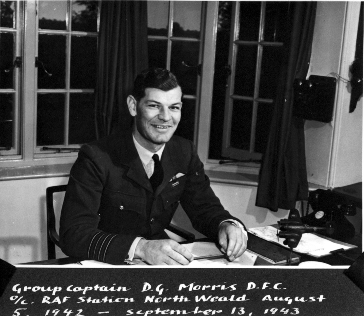 1 person i militæruniform bak et skrivebord.  Kaptein D. G. Morris D.F.C. Håndskrevet engelsk tekst under bildet.