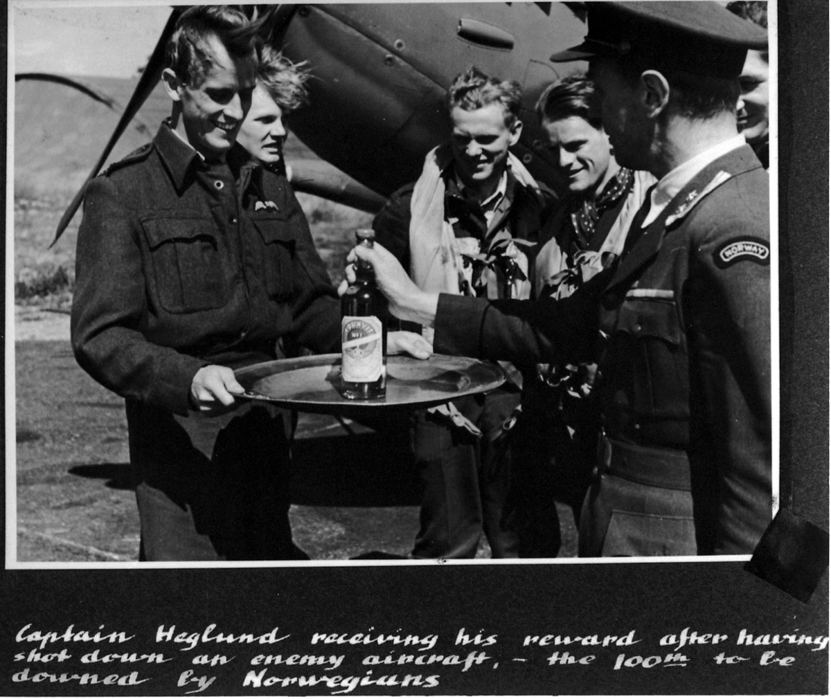 5 personer i militæruniform, 2 med pilotutstyr, foran fly. En av dem holder et brett med en flaske på. Engelsk håndskrevet tekst under bildet.