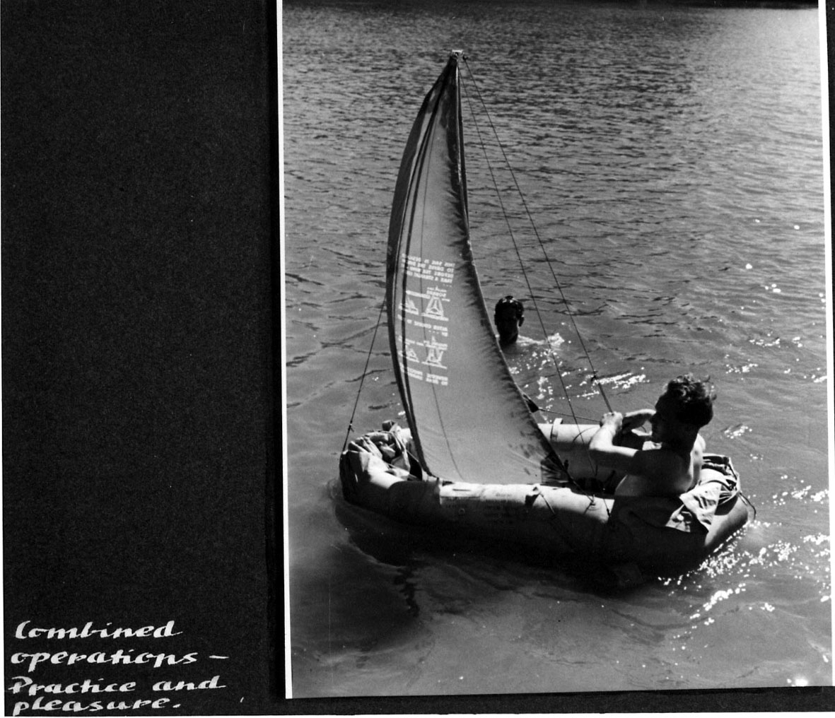 Gummiflåte med seil. En person ombord. Seiler bortover vannet. (Håndskrevet tekst under bildet.