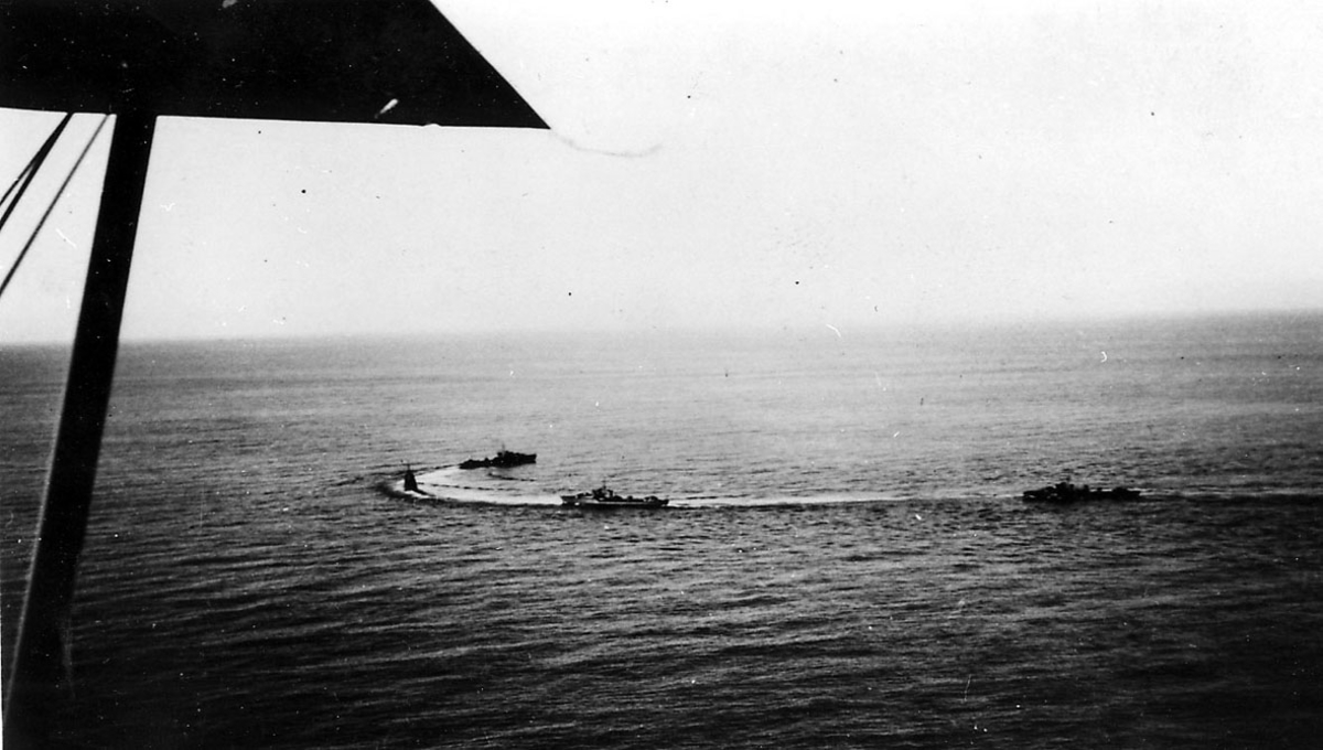 Luftfoto, litt av ene vingen på flyet bildet er tatt fra sees. Under sees 3 militærfartøy,  i fart på havoverflata.