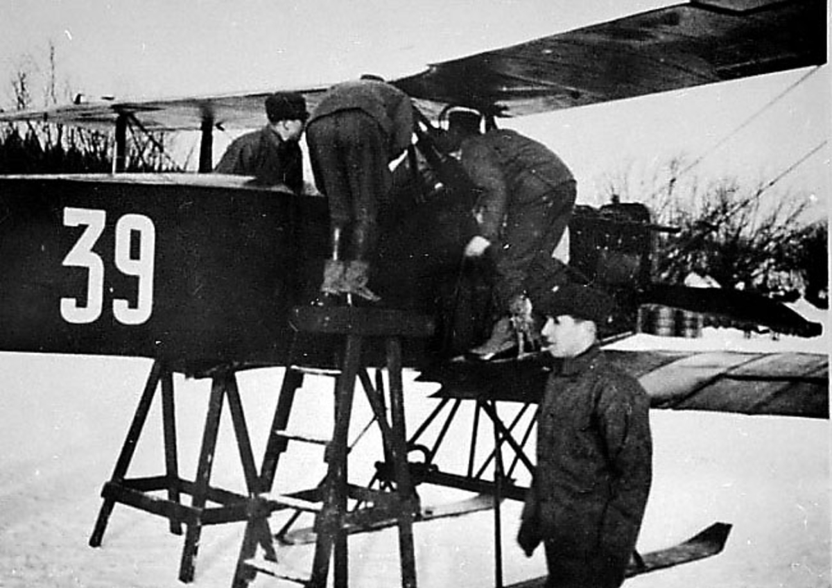 1 fly på bakken med skiunderstell, F. F. 6 "Kaje I" 39 fra Hærens Flyvåpen. Flere peroner ved flyet. Forpartiet skrått fra siden. Snø på bakken.