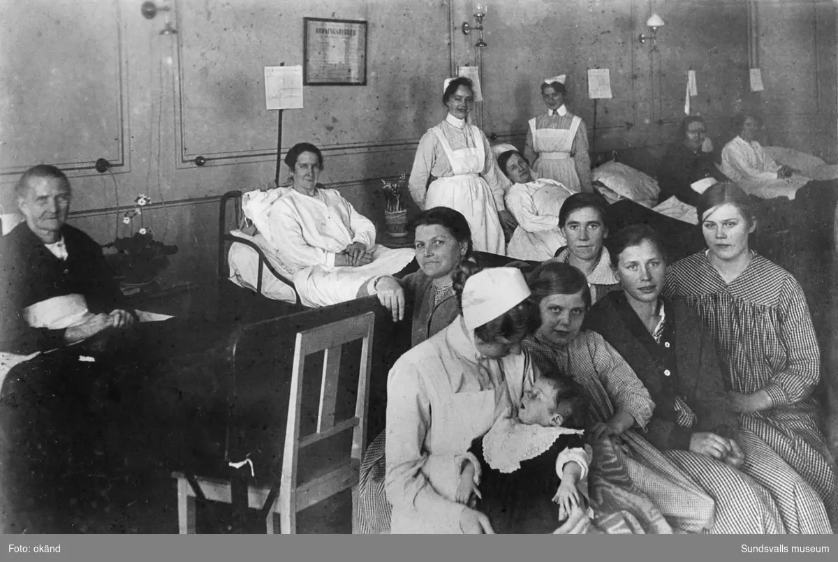 Sjukhussal på Sundsvalls lasarett. Frida Vikström (mitt i bilden) sitter i sängen. Sjukjournaler syns ovanför sängarna samt en tavla med ordningsregler på väggen.