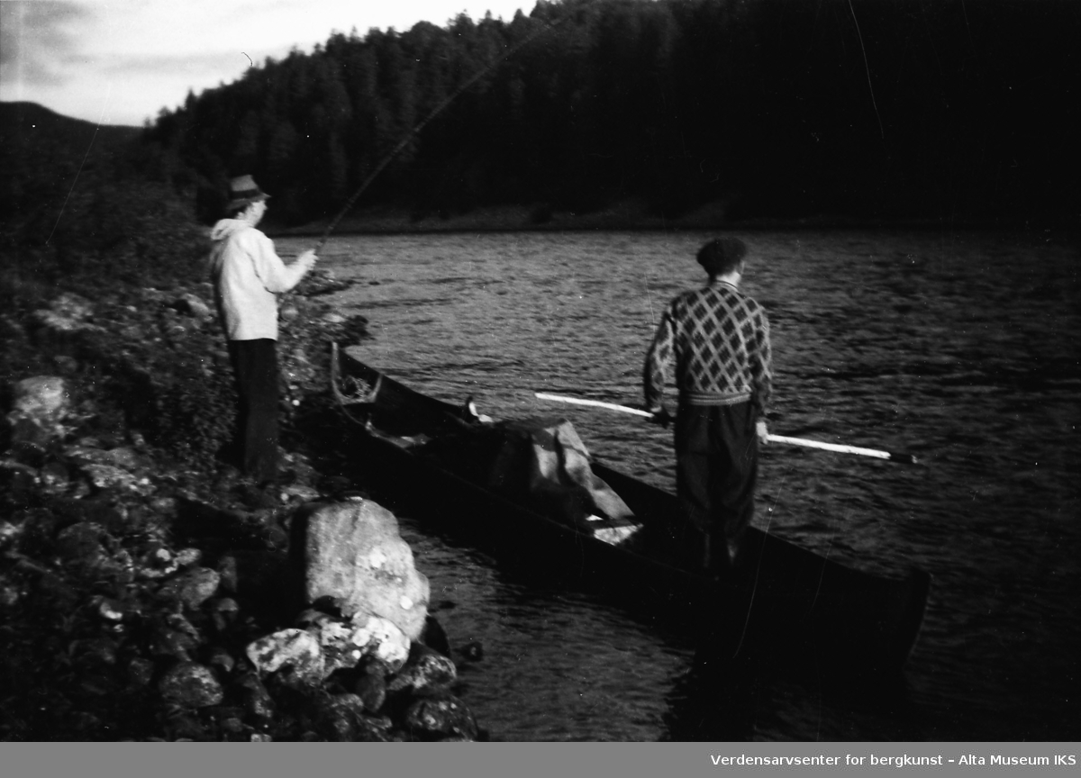 Ole Roald står med stanga på elvebredden, og en mann står klar i båten til å ta laksen.
Bildet er tatt i fiskesesongen på sommeren i 1949.