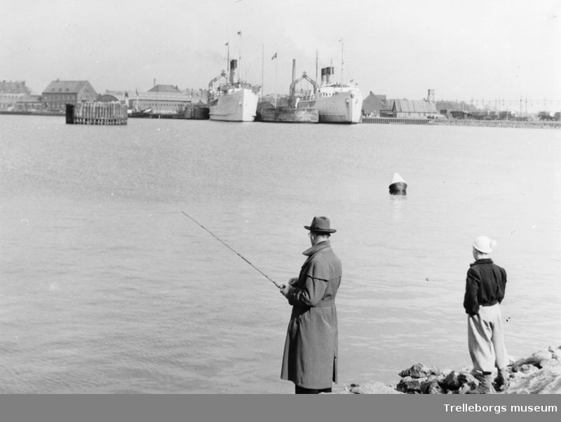 Manen i Trelleborg. På bilden står en man och fiskar i hamnen, pojken brevid tittar på färjerna.