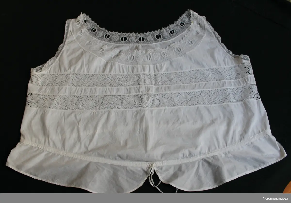 Liv/underskjorte for dame. Tynt bomullsstoff. Løpegang ved hoften.. Innfelte blondebånd over brystet og ved avslutning ved armer og hals. Maskinsøm.