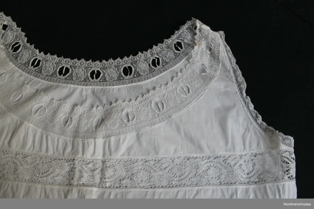 Liv/underskjorte for dame. Tynt bomullsstoff. Løpegang ved hoften.. Innfelte blondebånd over brystet og ved avslutning ved armer og hals. Maskinsøm.