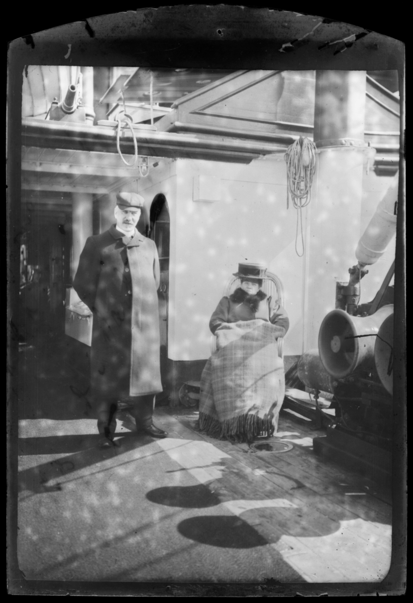 En kvinne sitter på dekkstol ombord på en hurtigrute god kledd og en ullteppe god rung seg. En mann står ved sidan av hun kledd i lang jakke og en lue.