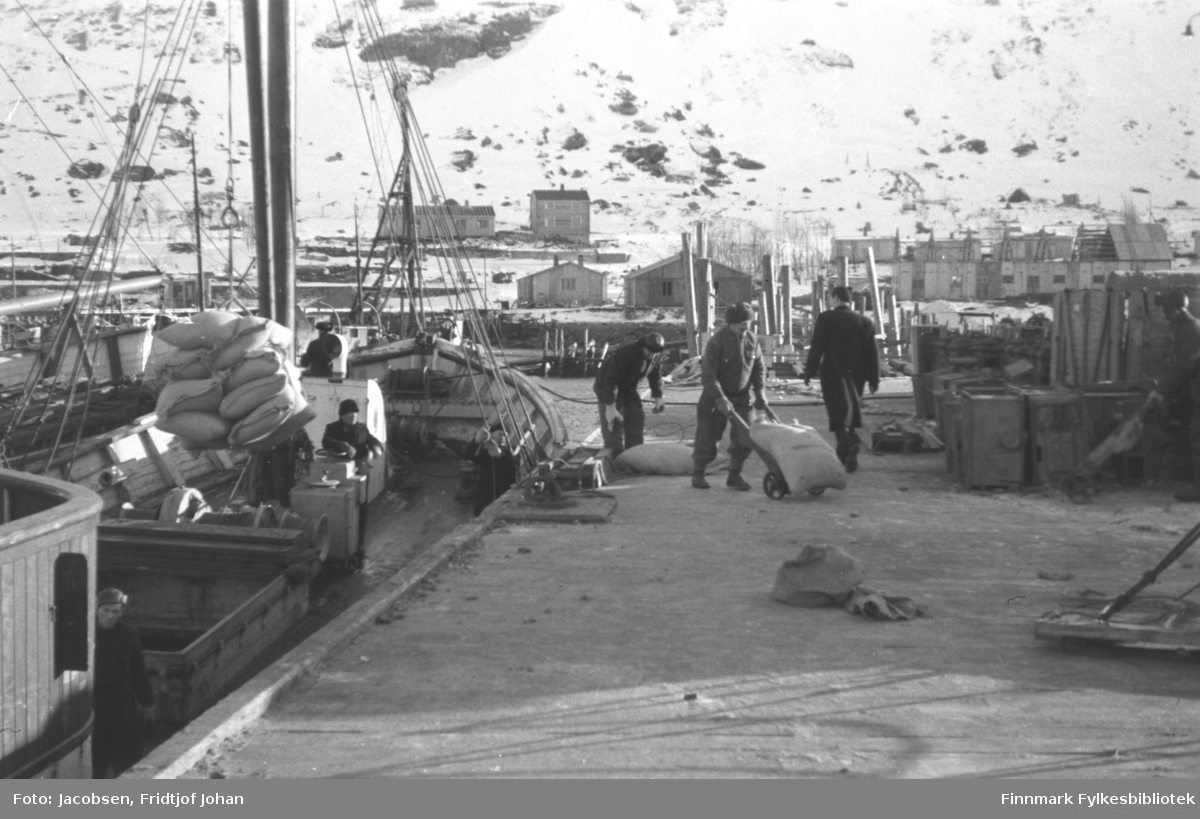 Nissenkaia i Hammerfest. Forsyninger strømmer inn vinteren 1945-46. Mannen som går med ryggen til er Ragnvald Jacobsen. Tre andre menn er i ferd med å losse fraktebåten som ligger ved kai. En fjerde står ved lasterommet, nede til venstre på bildet. Til høyre på bildet står endel trekasser og i bakgrunnen har noen brakker blitt satt opp og noen i ferd med å bli satt opp. Det snødekte fjellet i bakgrunnen er Salen