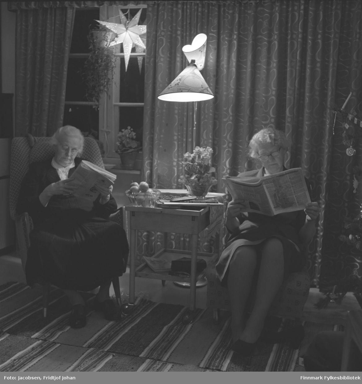 Jul i Storvannsveien hos Aase og Fridtjof Jacobsen. I lenestolen til venstre sitter Olga Jacobsen. Hun er iført en mørk kjole med hvit krage og leser en avis. I stolen til høyre sitter Ingeborg Jacobsen. Hun har også mørk kjole og leser en avis eller tidsskrift. Begge damene bruker briller. Mellom dem står et lite bord med en blomsterdekorasjon og et fruktfat oppå. På gulvet rett bak står en lampe og i vinduet står og henger det flere planter og en julestjerne. Ganske mørke og tykke gardiner henger delvis foran vinduet. To matteryer ligger på gulvet der de sitter.