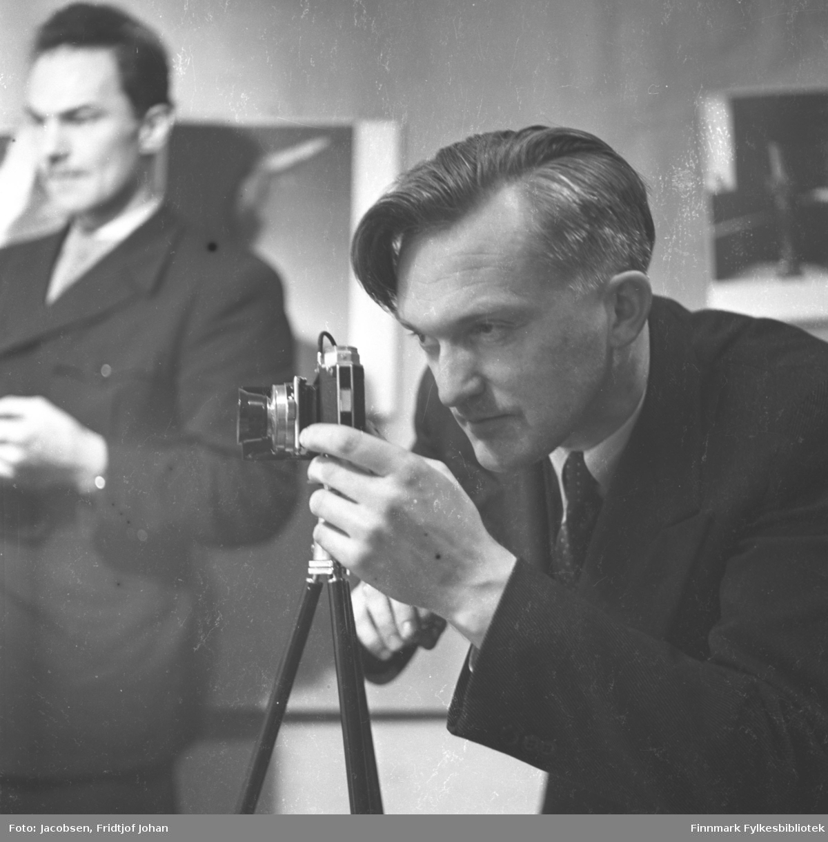 Fridtjof Jacobsen fotografert av Alf Edvard Jacobsen mens han selv fotograferer. Han har en mørk dress med hvit skjorte og slips på seg. Kameraet står på et stativ. En mann i mørk dress, hvit skjorte og slips står til venstre på bildet.