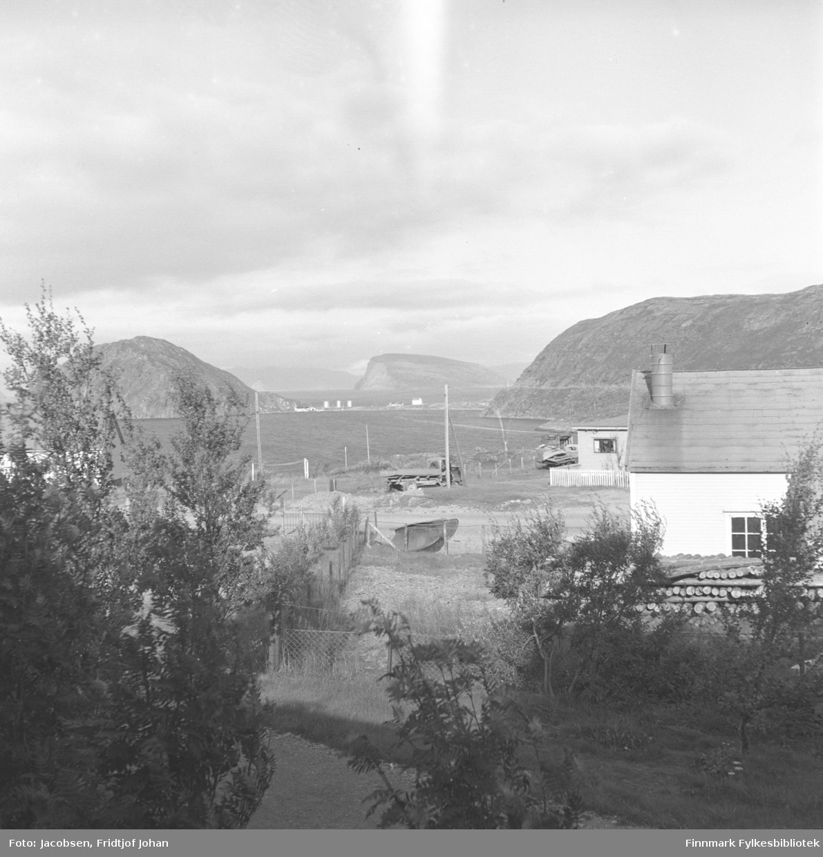 Utsikt fra Arne Nakkens tomt i Rypefjord. En god del løvtrær står i forgrunnen med et nettinggjerde rett bak. El-stolper står langs Fjordaveien, midt på bildet. Et lite hus ses bak Veivesenets brakke som står til høyre på bildet. En stor stabel med tømmerstokker ligger langs veggen. Over havna/Rypfjorden ses Essos tankanlegg på Rypeidet. Fjellene på sidene er Rypklubben til venstre på bildet og Rypefjell til høyre. Øya Håja ses ute i Sørøysundet rett over tankanlegget.