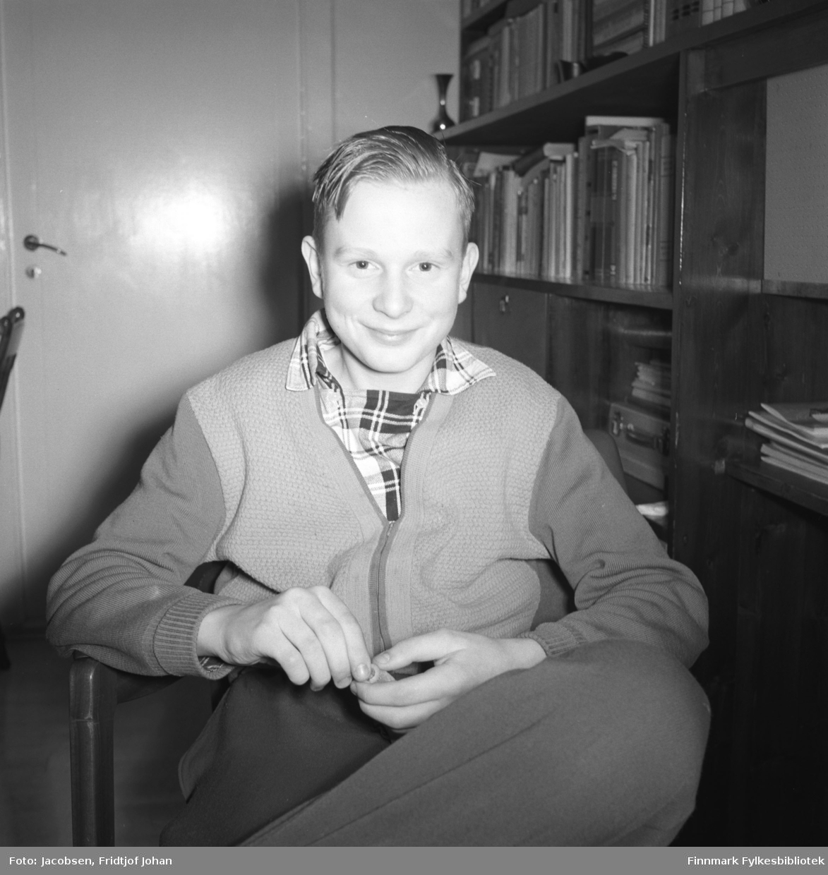 Arne Jacobsen poserer for fotografen. Han sitter i en lenestol iført en ganske mørk bukse, litt lysere genser/jakke og rutet skjorte under. En mørk veggseksjon står langs veggen til høyre på bildet. En del bøker står i hyllene og noen papirer/hefter ligger også der. Til venstre på bildet står en dør som er lukket.