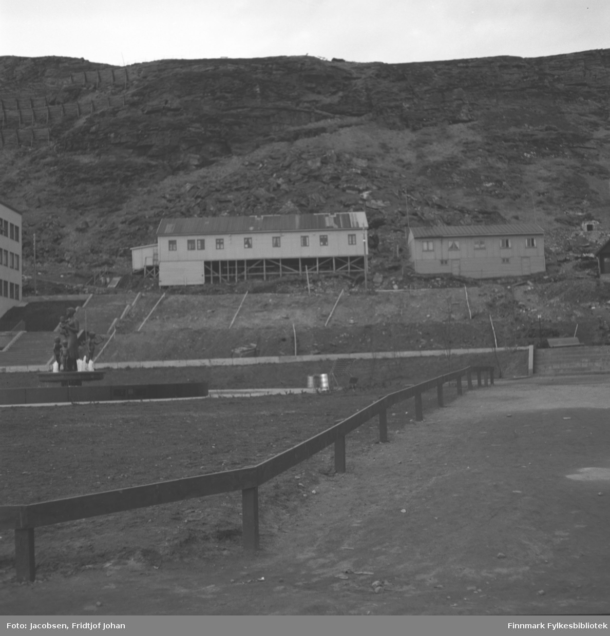 Rådhusplassen, fontenen på Ole Olesens plass til venstre i bildet. Bygningen som såvidt syns til venstre på bildet er DNB-bygget. Starten på sikksakkveien ligger like bak. Lengst til venstre i den lange brakka midt på bildet begynte Ernst Larsson sin sportsforretning i 1948. Dette var en av de siste brakkene som ble revet i Hammerfest, og den hadde nummer 276. I den høyre delen av brakka drev B. Hansen forretning. Fjellet i bakgrunnen er Salen og noen skredvern ses i fjellsiden.