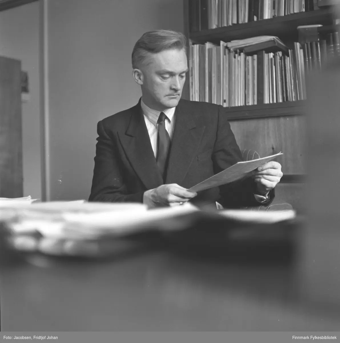 Fridtjof Jacobsen fotografert mens han leser et dokument. Sannsynligvis tatt på hans arbeidsplass ved et av Televerkets kontorer. Han har en mørk dressjakke, hvit skjorte med noen striper og mørkt slips på seg. En bokhylle står på på veggen bak han og den er full av bøker og hefter.