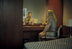 Aase Jacobsen på hotell. Hun sitter i en enkel stol foran et