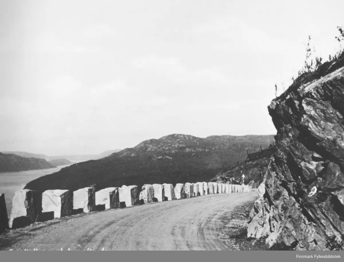 Postkort fra Storfjellet ved Langfjorden. På fotografiet som er tatt i en sving på veien, kan man se utover langfjorden og fjell i området. Langs grusveien på venstre side er det satt store steiner langsetter som et autovern. På øversiden av veien er det fjell