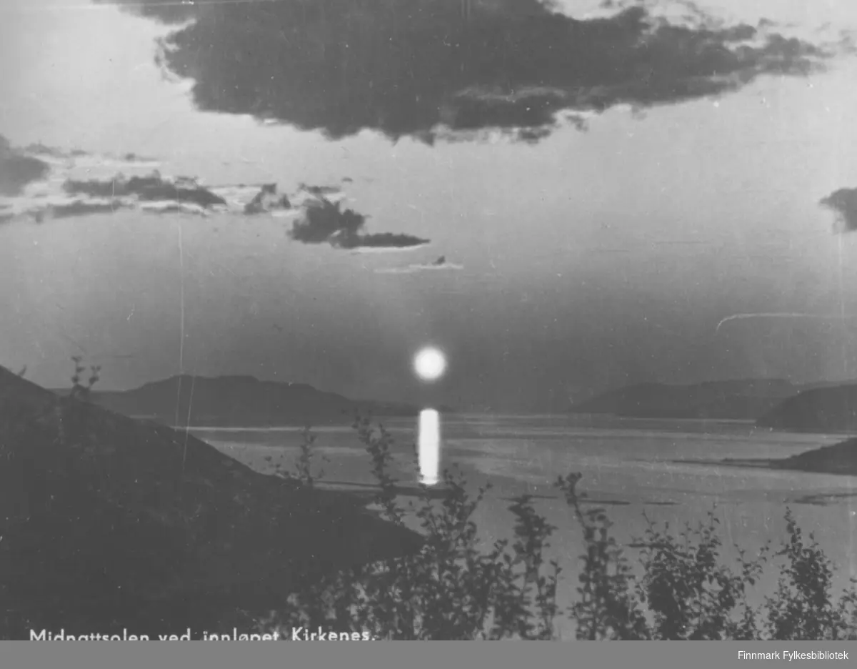 Postkort Fra Kirkenes. Midnattsola speiler seg i sjøen ved innløpet til Kirkenes. Vannet er omkranset med fjell. Det er noen mørke skyer på himmelen. I front av bildet kan man se trær