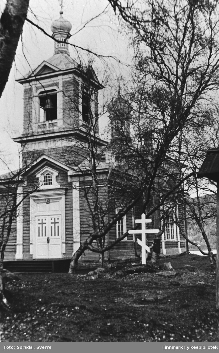 Bilde av kirken i Boris Gleb. Trekirke. Ser klokken i tårnet og kirkedøren mot oss. I forgrunnen står det et gravkors.