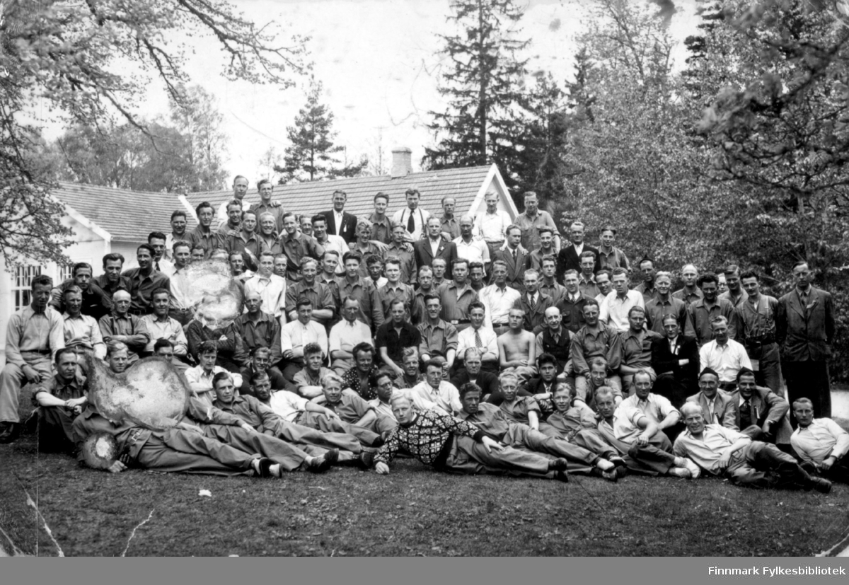 Fotografi av en stor gruppe menn som er hentet hjem med de hvite bussene ifra tysk fangenskap under andre verdenskrig. Magnus Stenbakk sitter i tredje rekke nedenfra, omtrent midt på bildet. Han er kledt i en mørk skjorte. Bildet er tatt ute på en gressplen. I bakgrunnen ses en del av en hvit bygning
