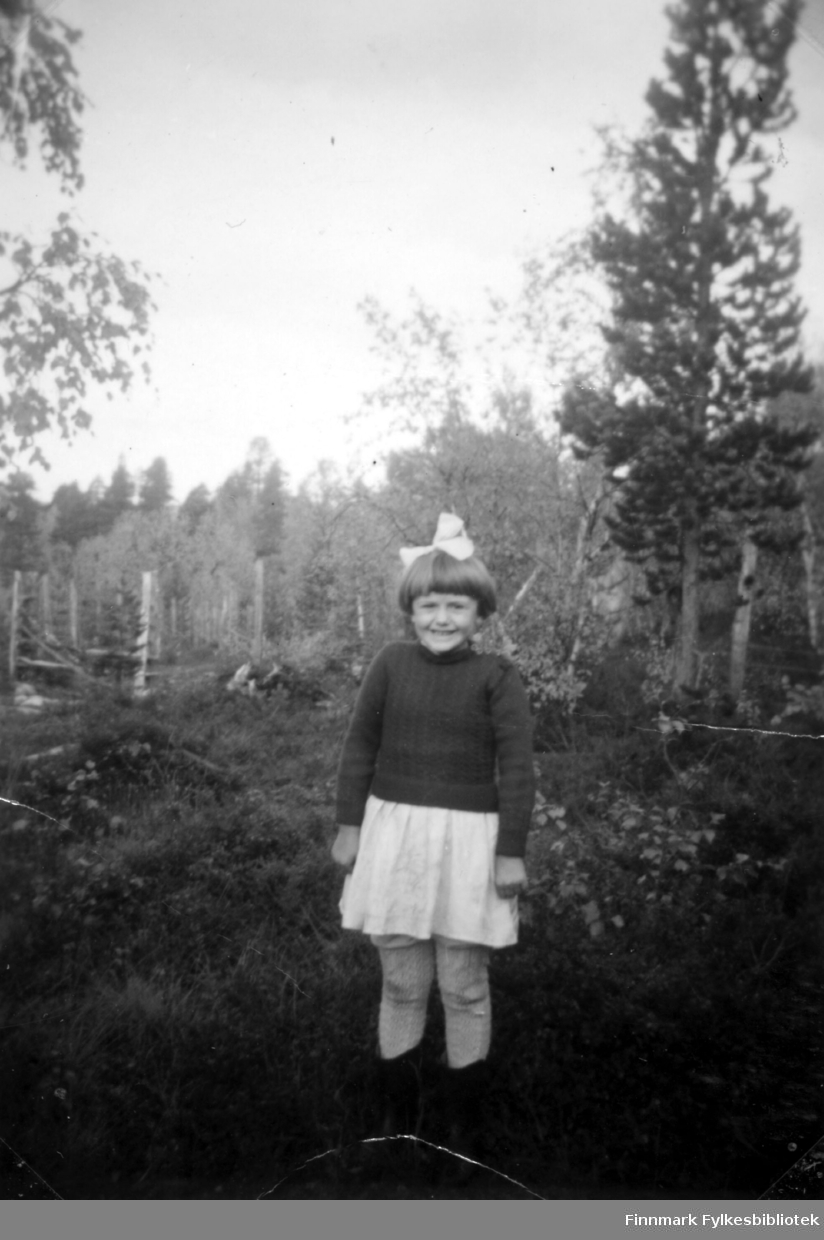 Fotografi av Edith Stenbakk søster til Agnes Randa. Bildet er tatt i bygda Namdalen i Pasvik. I bakgrunnen ser vi mange trær. Edith er kledt i en mørk genser og et hvitt skjørt. I håret har hun en hvit hårsløyfe