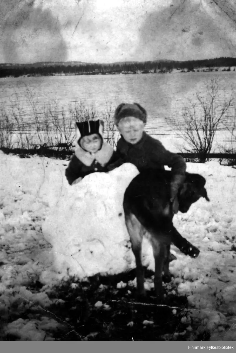 Fotografi av Mirjam og Eero Ranta. På bildet står de bak en stor snøklump. Det står en hund foran dem, som Eero holder armen sin på. Begge barna er kledt i luer og jakker. Mirjam har en krage rundt halsen. Det ligger snø på bakken i bakgrunnen ser man skog på andre siden av elven som er isfri