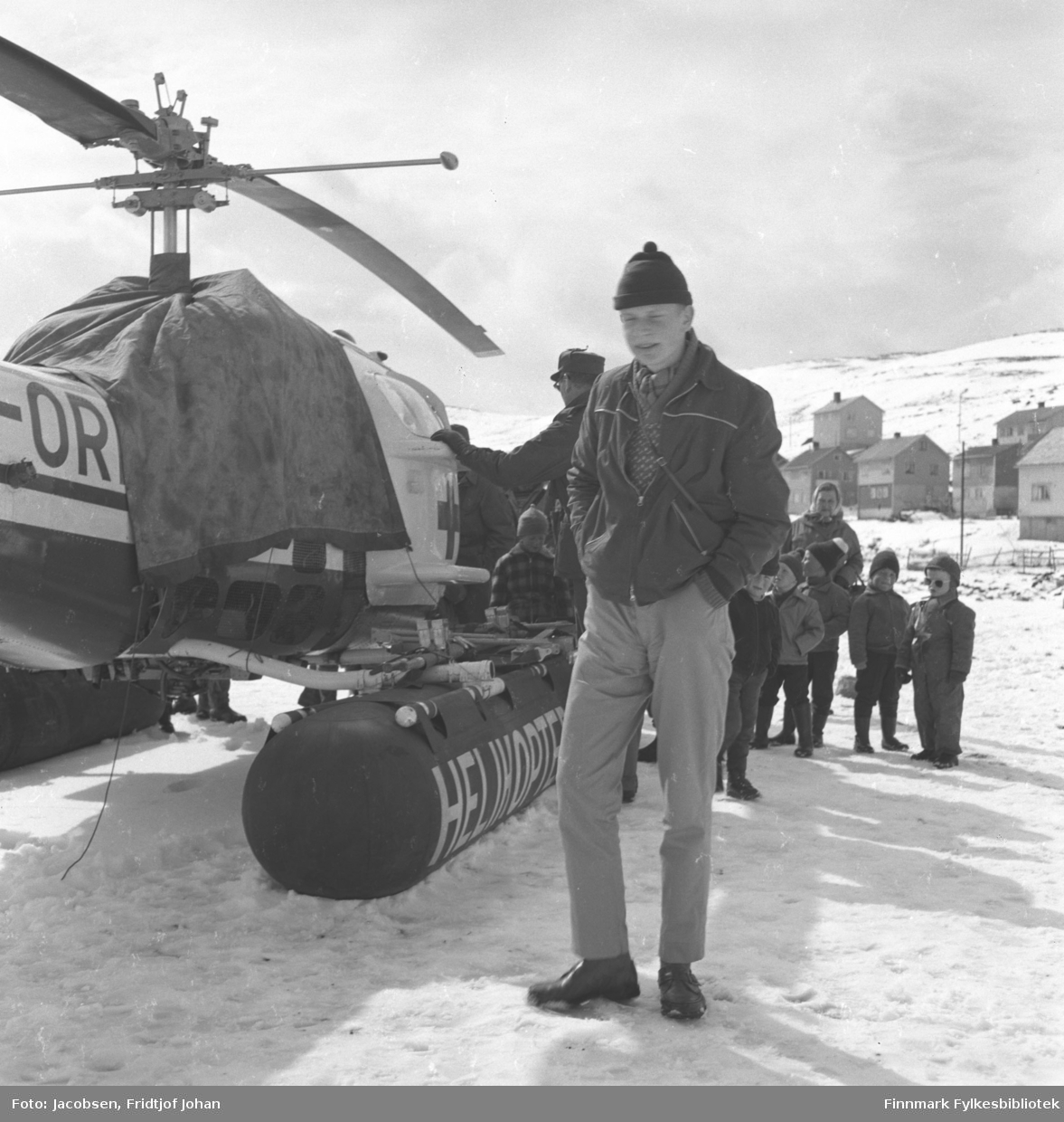 Arne Jacobsen står ved siden av et helikopter fra Helikopter Service A/S på Hammerfest stadion med noen barn bak han. Bebyggelsen i Skytterveien ses i bakgrunen.