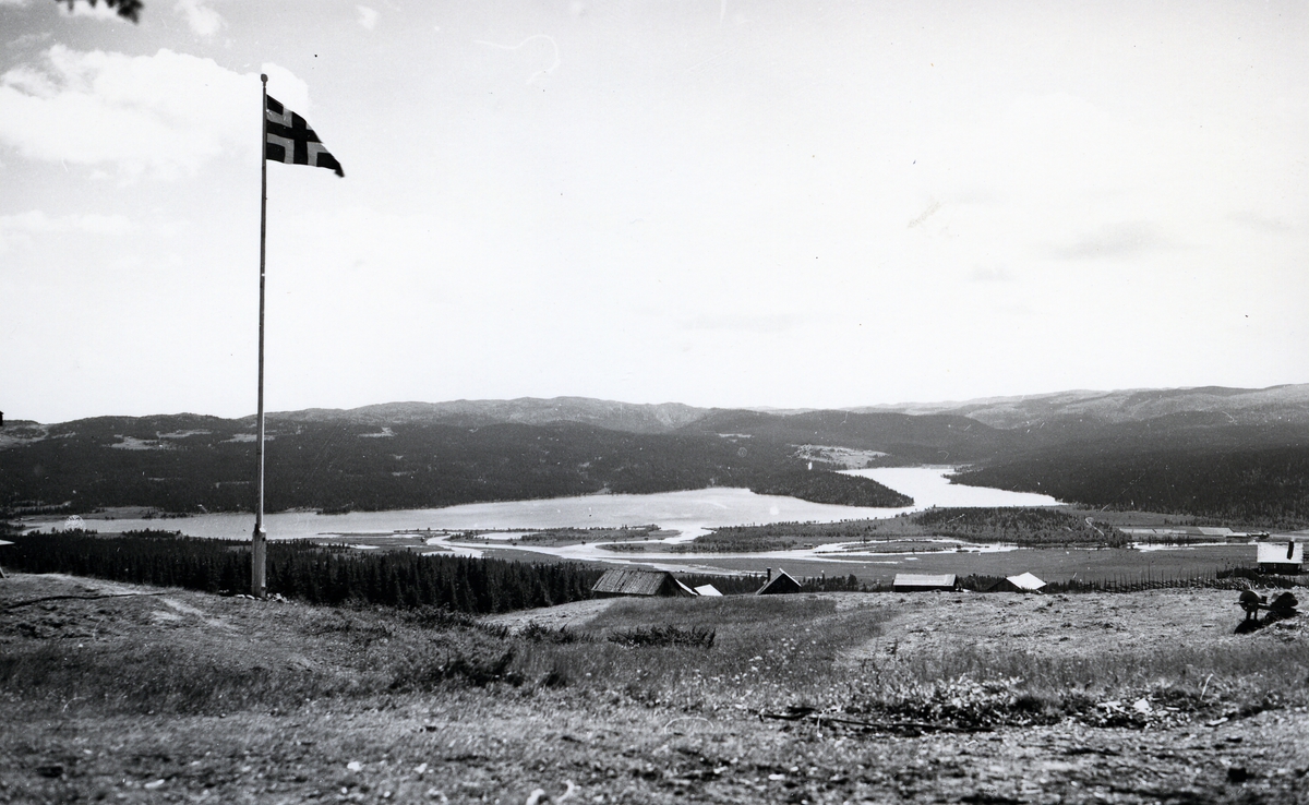 Utsikt over stølslandskap og innsjø, med flaggstang i framgrunnen