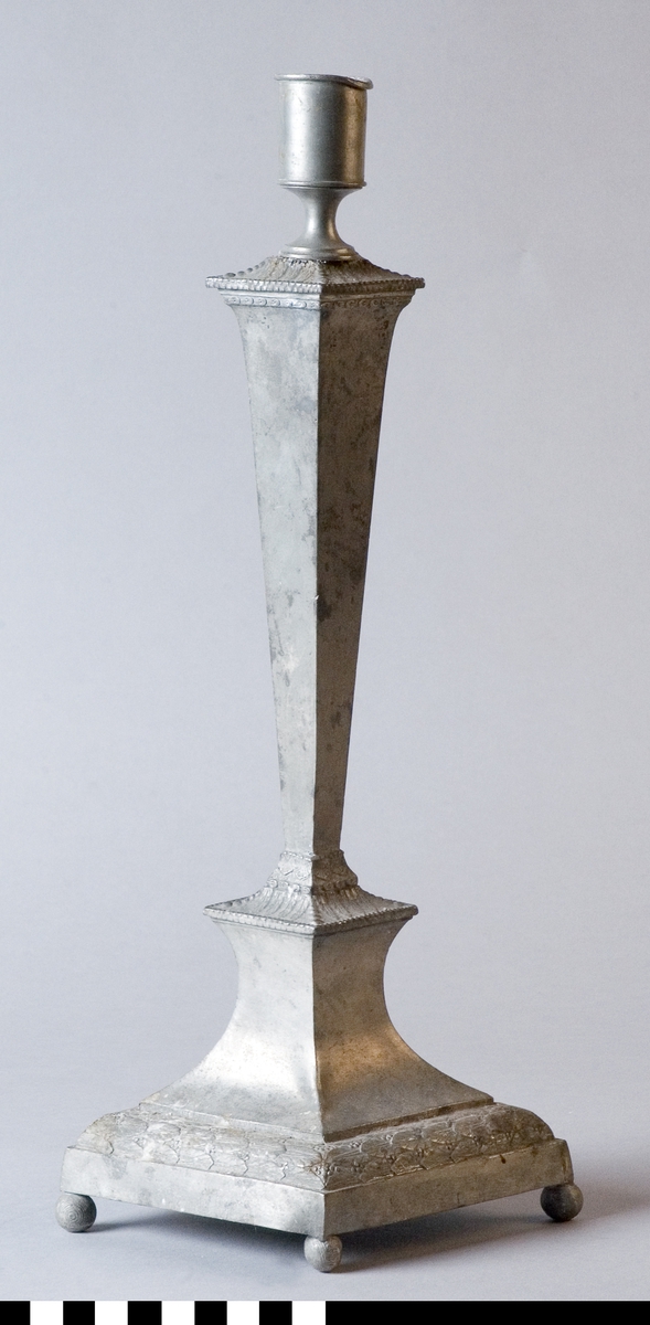 Ljusstake av tenn, möjligen engelskt tenn, britannica metall, med en ljuspipa. Fyrsidigt, utdragen rombiskt slätt ben med ljuspipa. Foten utdragen rombisk form, fyra kulfötter. Dekor av blandrankor i relief.