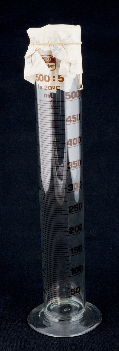 Målesylinder med stett og målestreker og tall fra 50 til 500 ml. Toppen er dekket med "apotekpapir" med strikk for å beskytte mot støv.