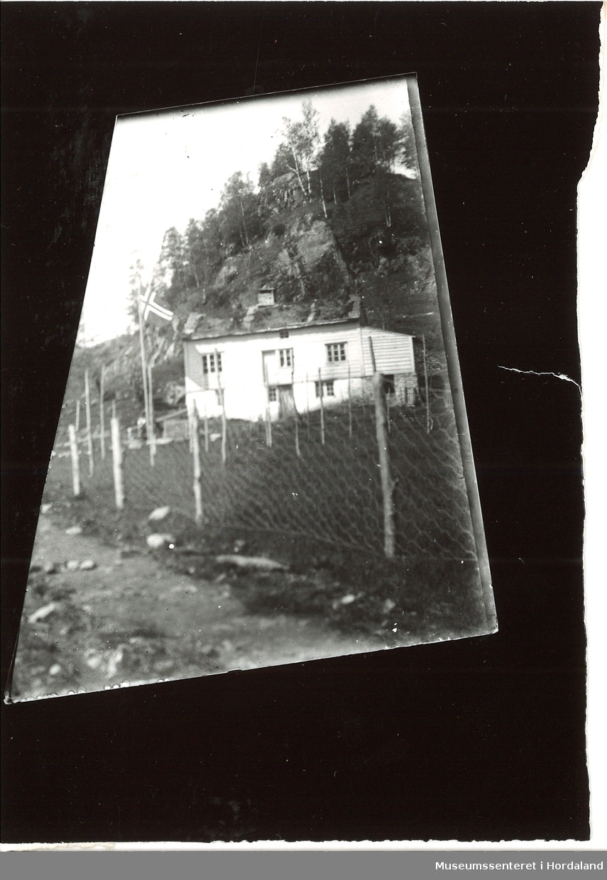 Bilde av et hus i Fossdal, med Norgesflagget heist, og hønsenetting-gjerde. Fjell og trær bak huset.