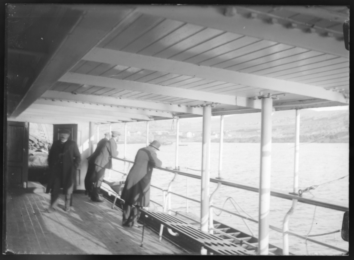 Ombord på hurtigruta under dekk, fire menn står ved rekka og ser mot land ved Makkaur. Vi ser også litt av interiøret ombord i hurtigruta i form av benker o.l
