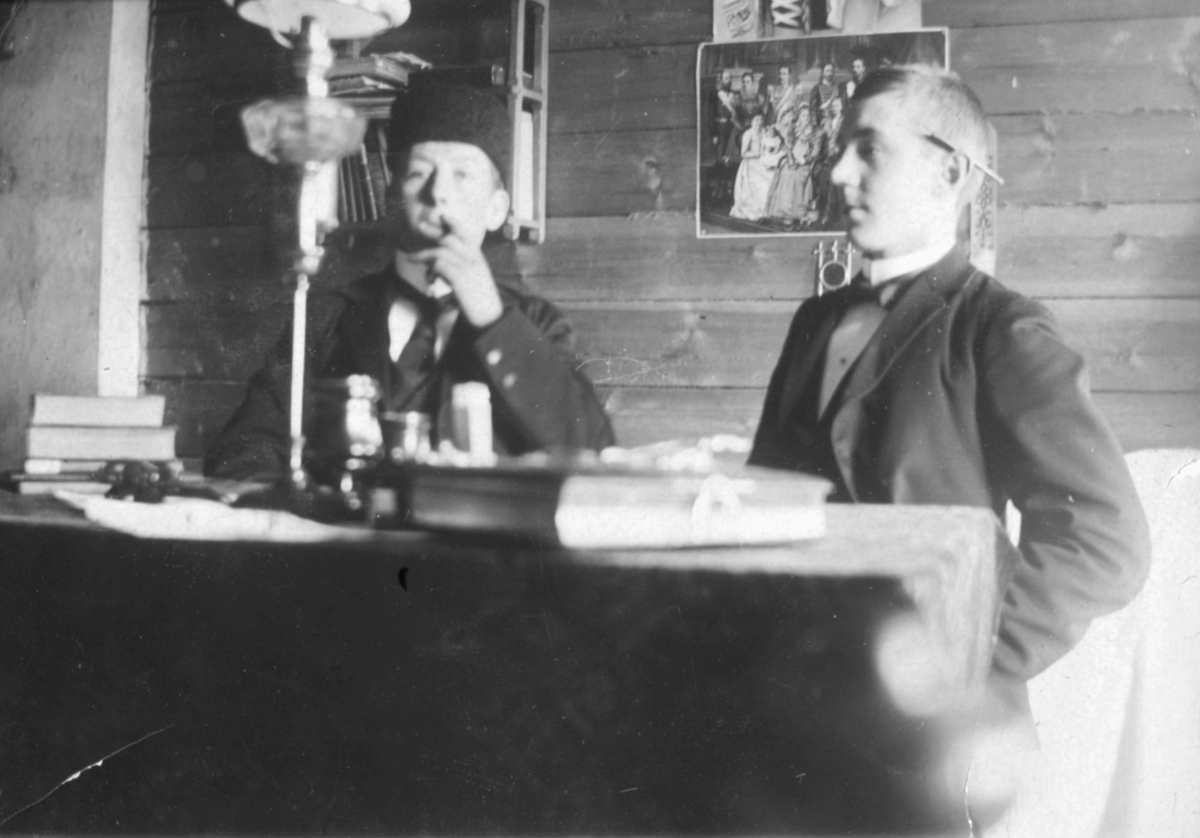 Dette bildet er trolig tatt på begynnelsen av 1900-tallet. To unge menn sitter ved et bord. De er kledd i jakker og vester. Mannen til venstre har på seg slips og lue. Han ser ut til å røyke. Mannen til høyre har en penn eller noe lignende bak øret og en sløyfe rundt halsen. På bordet kan man se bøker, papir og en lampe. På veggen bak kan man se fotografier av kvinner og en bokhylle.