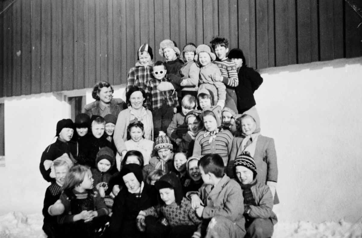 Gruppebilde av 1. og 2. klasse i Seida skole i 1955. Barn står foran skoleveggen i en fin vinterdag. En del av jentene har kledd seg i genserer, de fleste av guttene har på seg en jakke. Nesten alle har en lue på seg. To lærerinner står med elevene. Vi vet ikke navn til elevene eller lærerinnene.